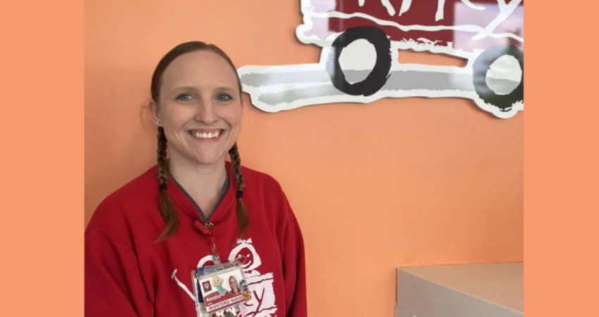 Kendra Kirby, a registered nurse, wears red scrubs and stands in front of the Riley sign at IU Health Fishers Pediatric Emergency Medicine department.